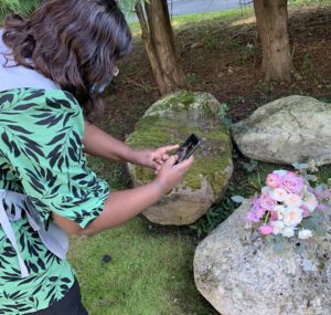 The student is learning how to take photos of her work with a cell phone during a floral workshop under a shaded area in the afternoon.